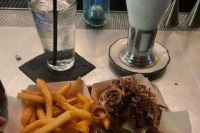 A delicious tray of fries and a burger topped with crispy onions, served alongside a glass of water and an over-the-top milkshake with whipped cream and chocolate drizzle at Black Tap Craft Burgers & Beer - 35th St