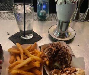 A delicious tray of fries and a burger topped with crispy onions, served alongside a glass of water and an over-the-top milkshake with whipped cream and chocolate drizzle at Black Tap Craft Burgers & Beer - 35th St